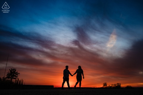In the glowing Kansas City sunset, a silhouette of a couple in love strolls hand in hand, the vibrant sky creating a stunning backdrop before their upcoming marriage.