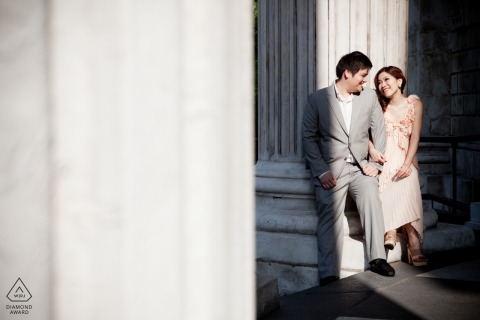 At the top of steps near massive old world stone pillars in the sunshine, a couple in London, UK posed for their engagement portrait session before their upcoming wedding.