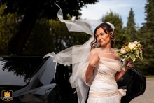 In Gorizia, Italy, a bride is gracefully exiting a black car on her wedding day, her veil elegantly caught by the wind, adding a touch of sophistication and style to this glamorous moment.